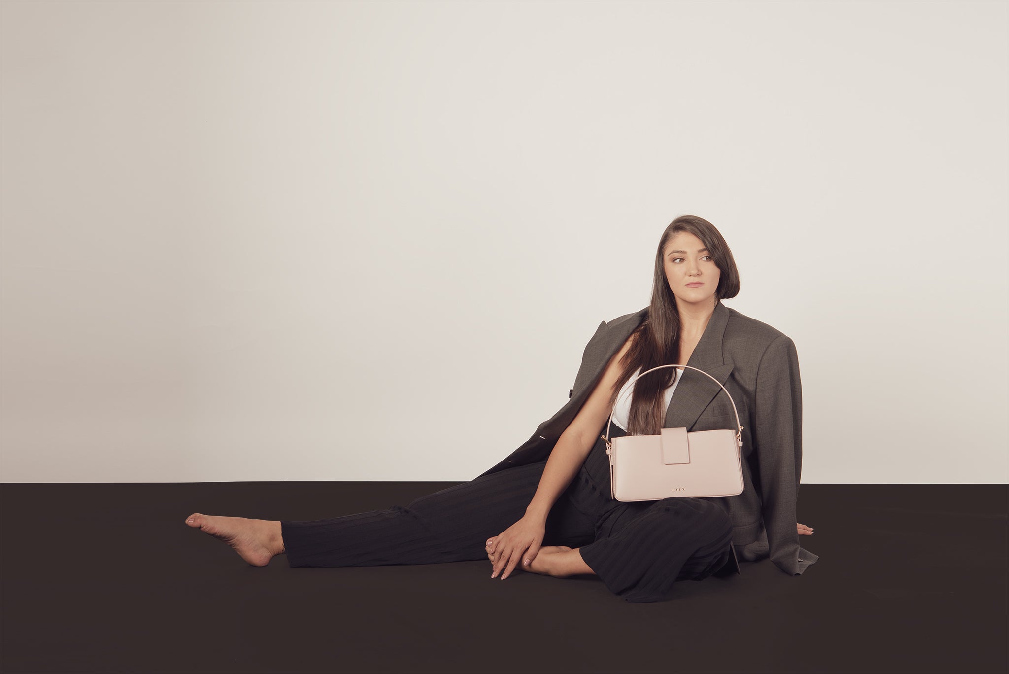 Woman sitting on a black floor holding a beige Belona handbag against a white background