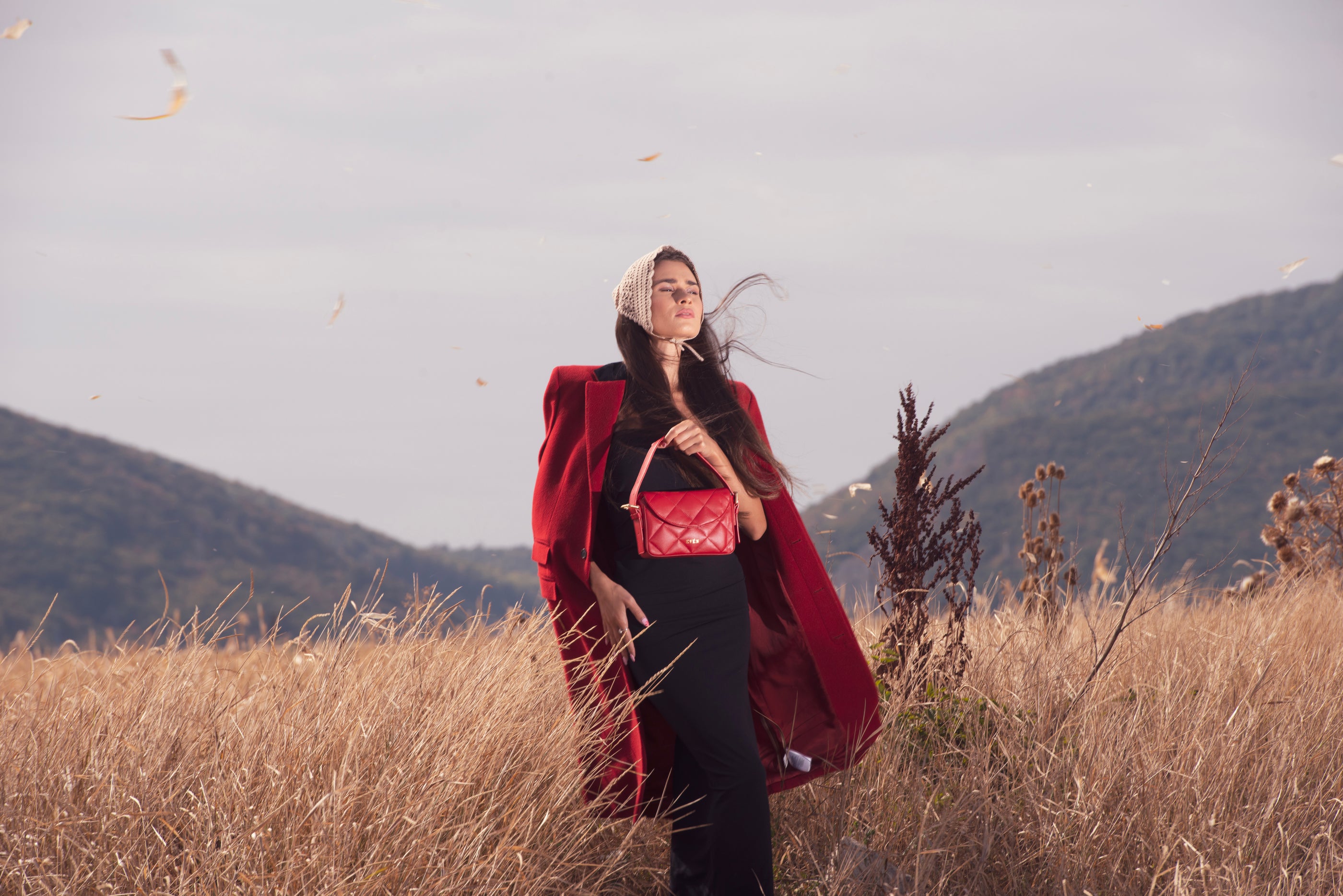 Woman in a red coat and black outfit standing in a field with mountains in the background