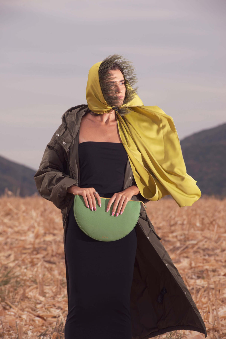 Person wearing a yellow hooded coat and holding a green clutch in a field with mountains in the background