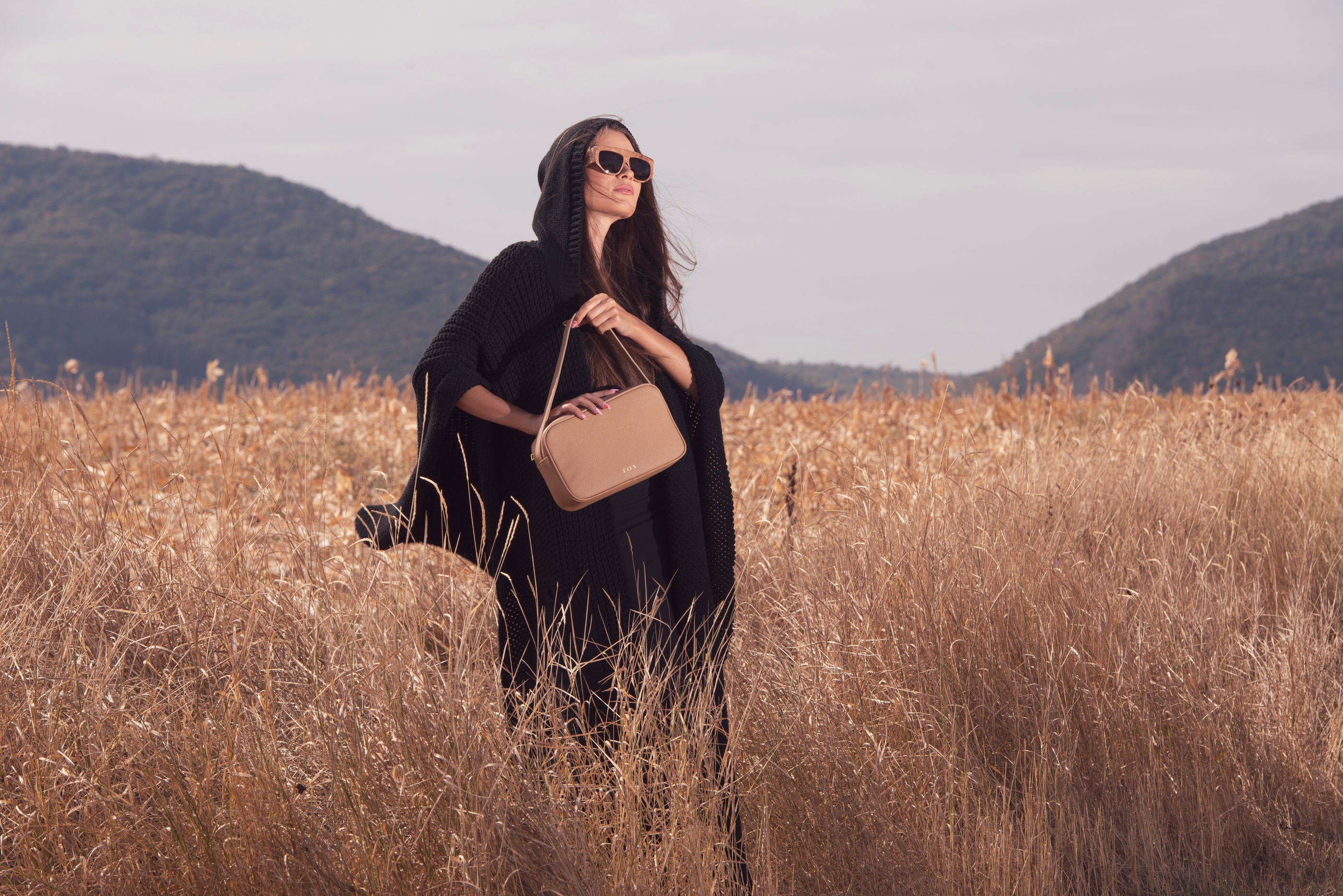 Woman holding a beige handbag in a field with mountains in the background