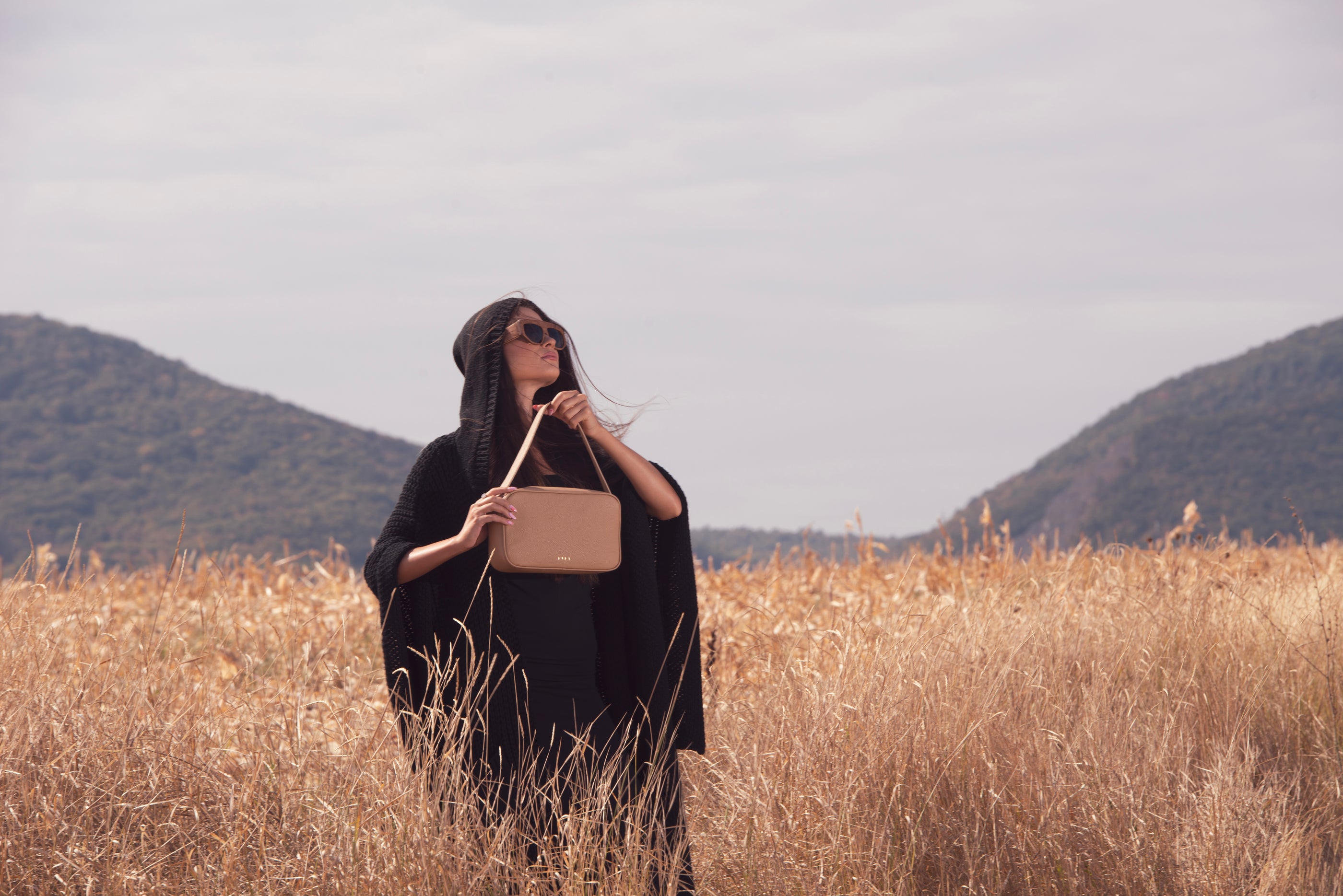 Woman holding a mocha color handbag in a field with mountains in the background
