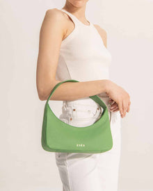 Lifestyle shot of Zoe Mini Baguette Bag in green leather with a sleek rectangular design, short strap for shoulder or hand carry, and minimalist gold-tone zipper, displayed on a white background. 
